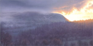 This is a signed limited edition print of an original soft pastel painting by artist Michael Howley. The subject of the painting is a beautiful dark, brooding image of mist over Pendle as the sun sets to the right. The dominant form of the hill is coloured in blue-greys and grey-greens and the clouds above are dark indigo. The misty forest in the foreground is in low-key dark purple-grey. There is a contrasting warm glow on the right hand side lighting the edge of the clouds and tree tops. The whole atmosphere of the painting seems to reflect the dark history of Pendle Hill and the Pendle witches.
