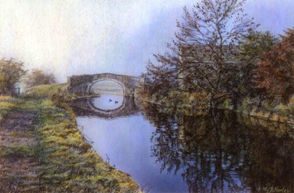 This is a signed limited edition print of an original soft pastel painting by artist Michael Howley. The subject of the painting is a scene of a bridge over the canal with a barn and autumn trees to the right. It is bathed in a gentle early morning light and the clear still blue reflections contrast with the autumn colours of the grassy bank.